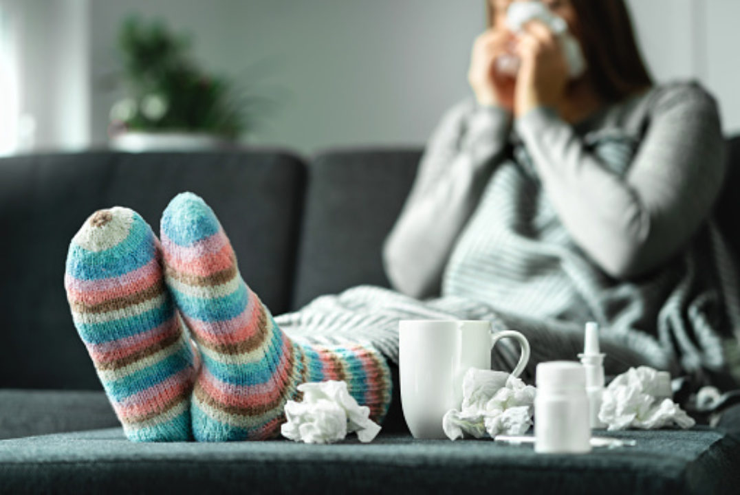 Sick woman with flu, cold, fever and cough sitting on couch at home wearing wool sock surrounding by tissue